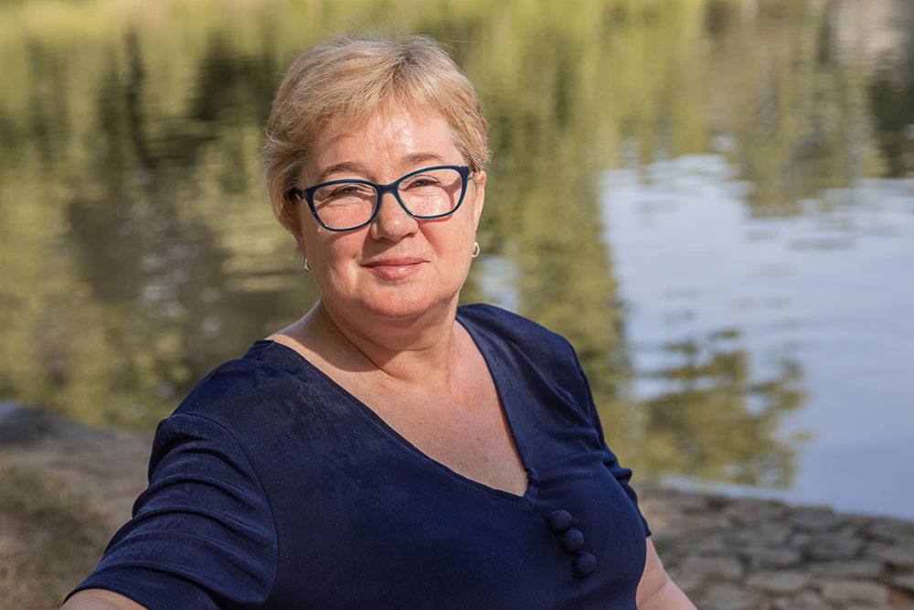 Natural Pharmaceuticals Movisan™ Collagen II Középkorú nő szemüvegben kék felsőben, mosolyogva a tóparton, természetes fényben.